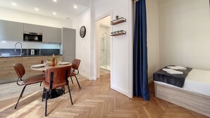 Et moderne køkken med indbyggede hårde hvidevarer, et spiseområde med parketgulv i fiskebensmønster, og en hyggelig sovezone med seng og lille bord.