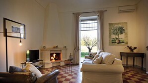 A living room with a fireplace, a sofa, a television, and a window with curtains. - Tenuta La Moresca – Villa with pool, and garden among the olive trees in Noto (Noto)