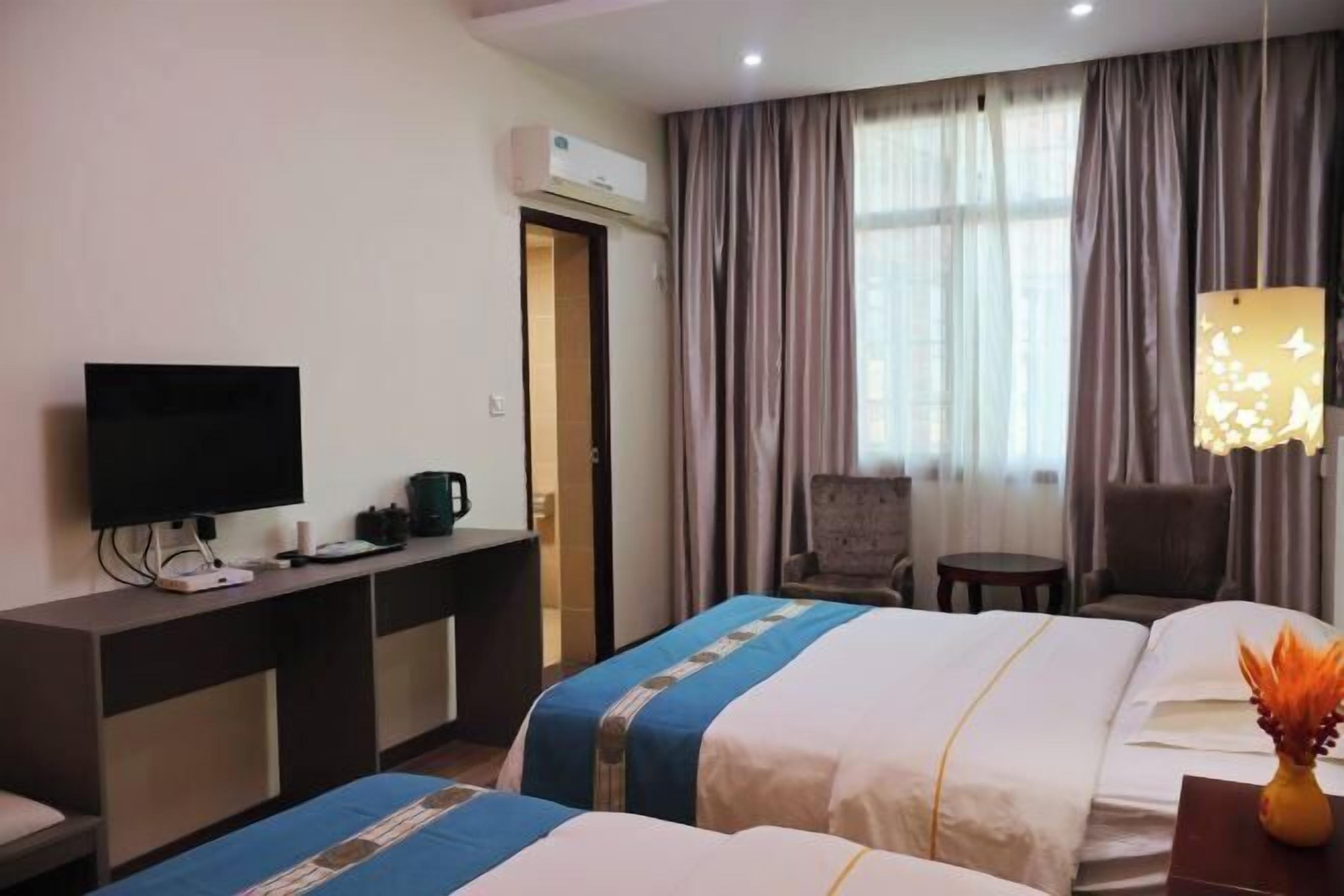 Habitación de hotel con dos camas, un escritorio con televisor, aire acondicionado, una silla, una mesita pequeña y una lámpara colgante decorativa.