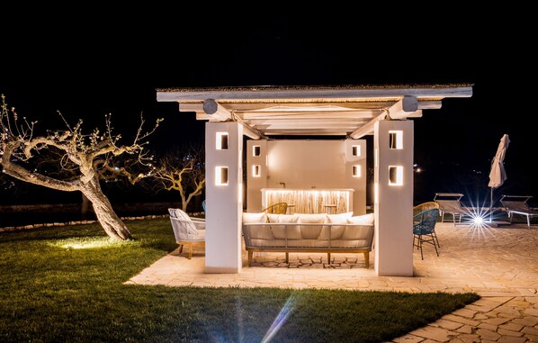 A covered outdoor seating area with a sofa and chairs, a pergola structure, and a tree in the background. - Elegant trulli and lamia for exclusive use, with heated pool. (Putignano)