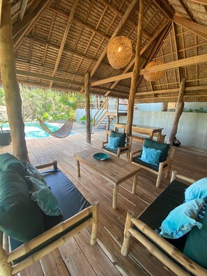 A wooden deck with a thatched roof, outdoor seating, and a view of a pool and greenery. - Villas Sampaguita (El Nido)