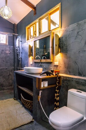 A bathroom with a stone wall, a wooden vanity, a white sink, a toilet, and a mirror with a wooden frame. - Villas Sampaguita (El Nido)