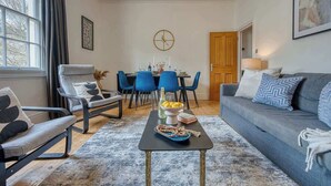 Een woonkamer met een bank, twee fauteuils, een eettafel met stoelen, een klok en een deur.