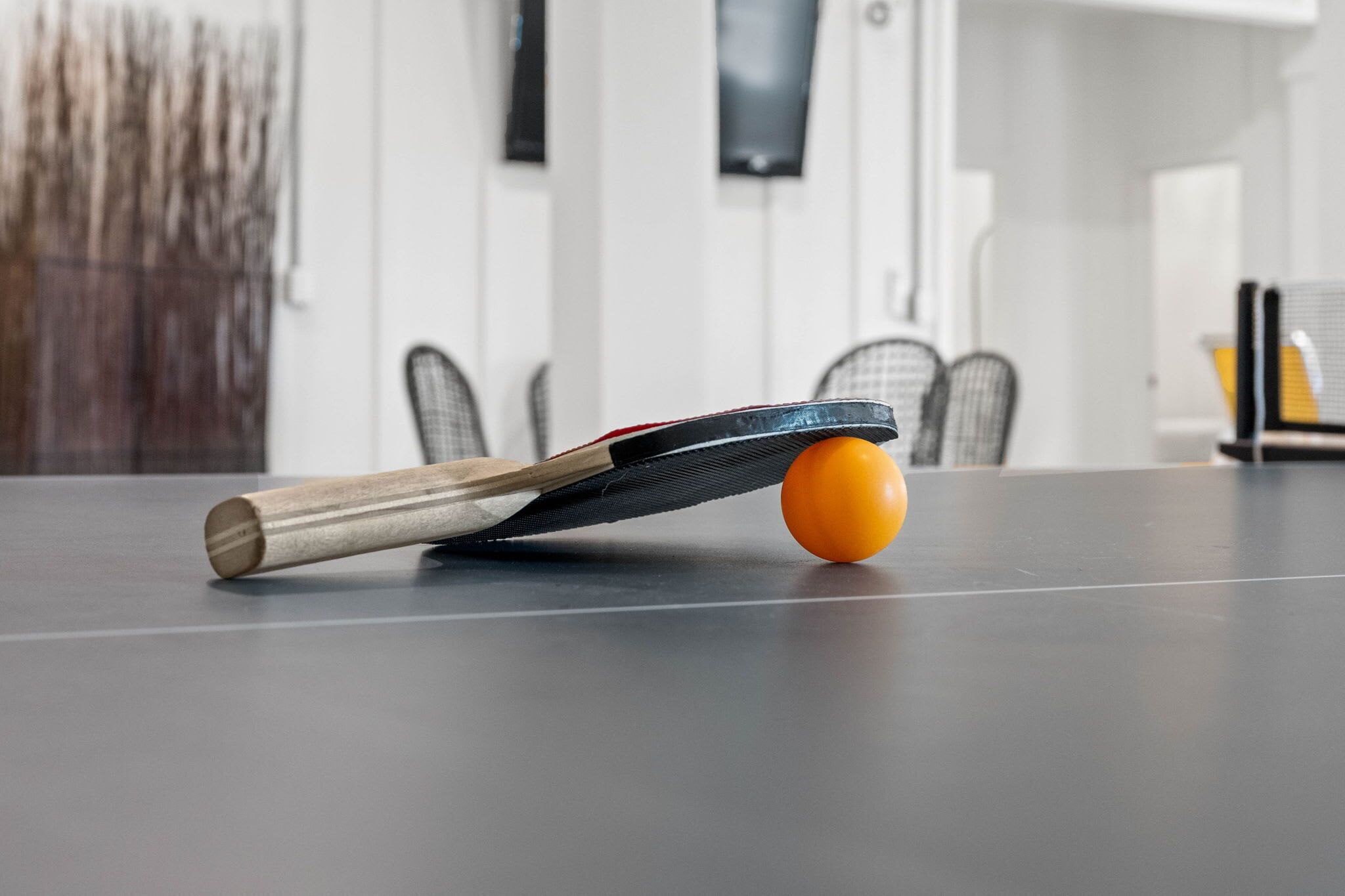 Ein Tischtennis-Tisch mit Schläger und Ball, Stühle ringsherum und ein Fernseher an der Wand.