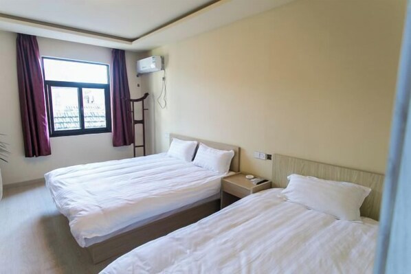 A hotel room with two single beds, a window with curtains, an air conditioning unit, and a bedside table with a lamp.