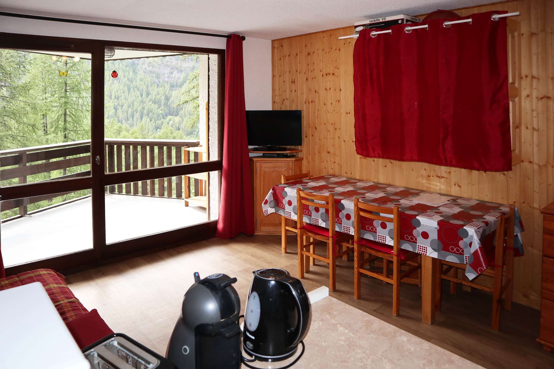A room with a balcony, a table with a tablecloth, a television, and two kettles.