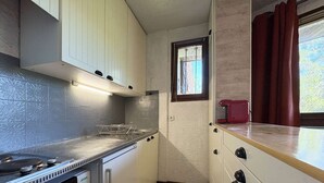 A compact kitchen with white cabinets, a stove, and a window with curtains. - 12 LA Seille Studio 4 Personnes (LES ORRES 1650)