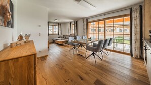 Ein geräumiger Wohnbereich mit Holzboden, einer Couch, einem Couchtisch und einem Esstisch mit Stühlen. Große Fenster bieten einen Ausblick nach draußen.