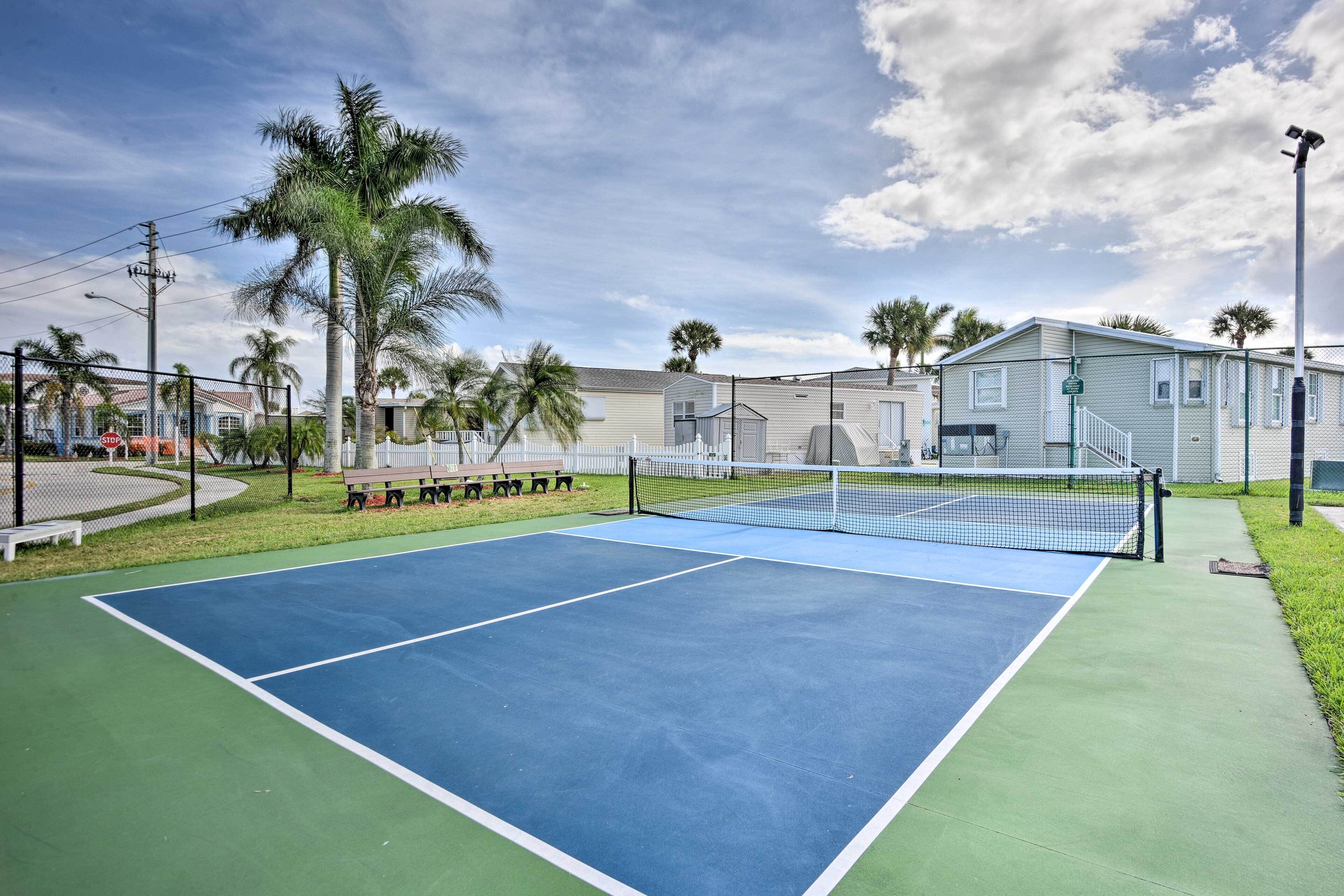 Ein Tennisplatz mit Netz, umgeben von einem Zaun, Palmen und einem Gebäude im Hintergrund.
