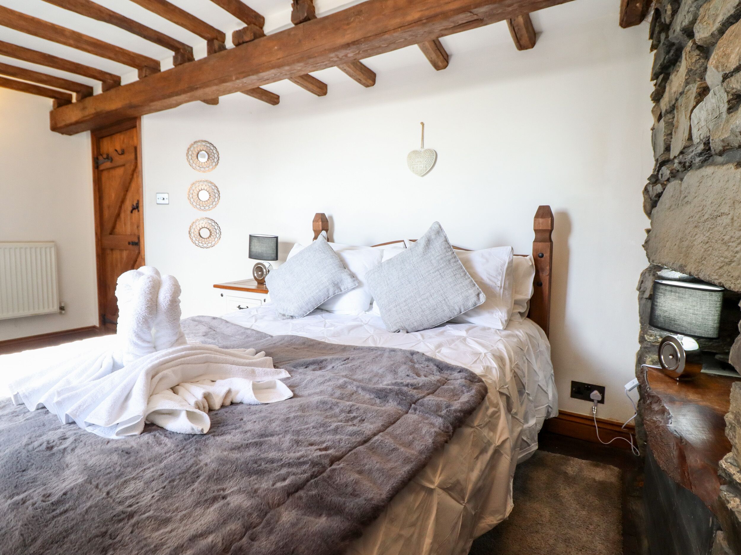 A cozy bedroom with a wooden headboard, a stone wall, and a bed with a grey blanket and pillows.