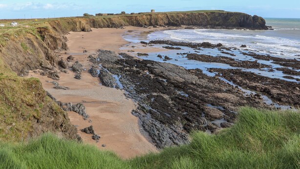 A coastal landscape with a sandy beach, rocky shoreline, and grassy cliffs.