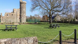 A historic stone castle with a tall tower, surrounded by a grassy area with picnic tables and a tree.