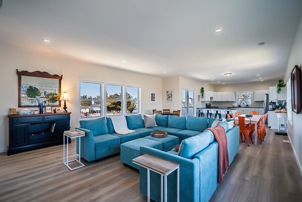 A modern living room with a sectional sofa, a small side table, and a dining area with orange chairs. - Lagoon Home with Dock + Bar -Near Calm, Kid-Friendly Beach (Little Egg Harbor Township)