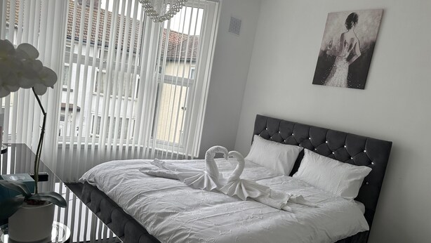 A neatly made bed with a white duvet and pillows, a dark headboard, and a wall-mounted painting. - House - Sleeps 6 - Garden - Seaside - Pet Friendly (Great Yarmouth)