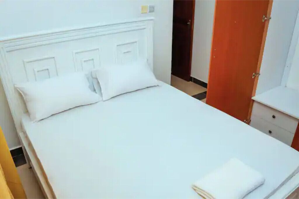 A neatly made bed with white linens and pillows, a wooden door, and a white dresser with drawers.