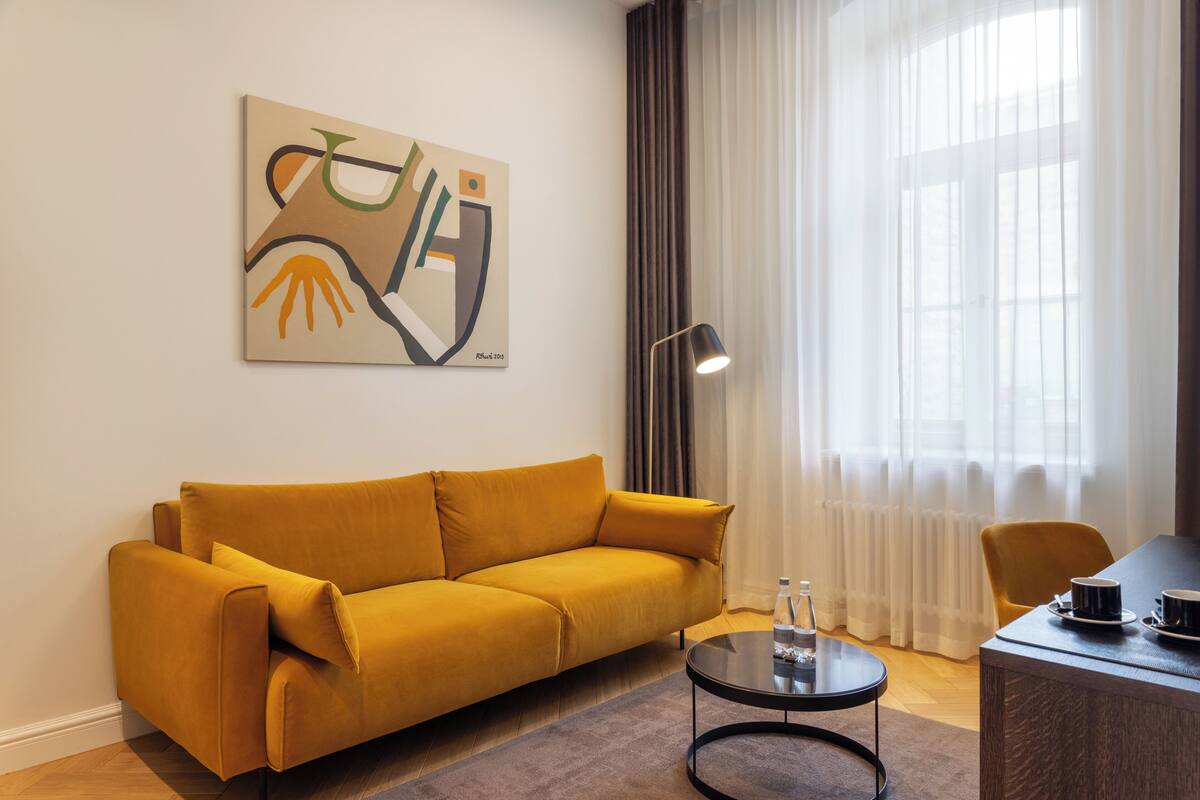 Een moderne woonkamer met een mosterdgele bank, een ronde salontafel, een schilderij aan de muur, een lamp en uitzicht door dunne gordijnen.