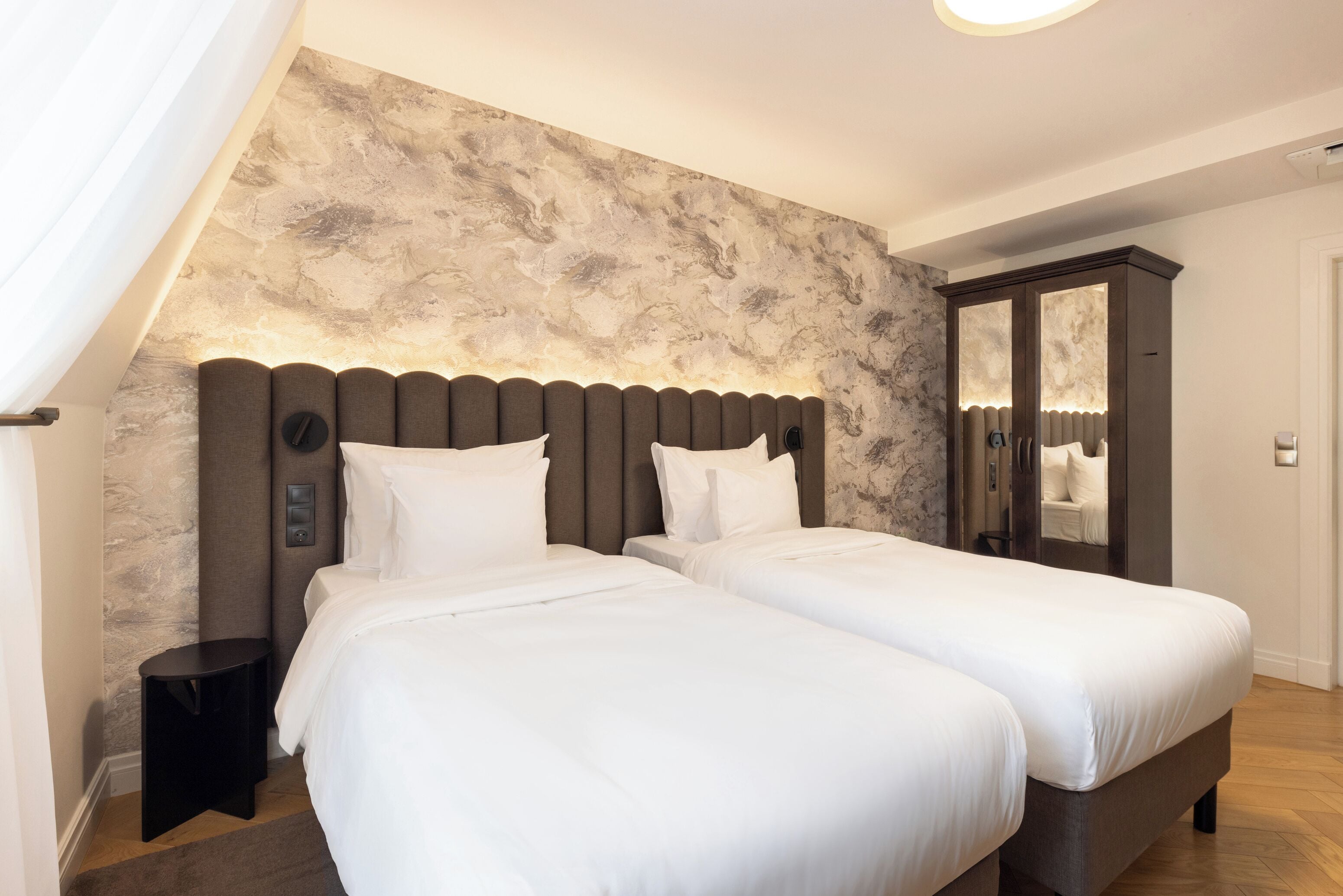 A modern hotel room with two beds, a marble-patterned wall, a dark headboard, and a large mirror cabinet.
