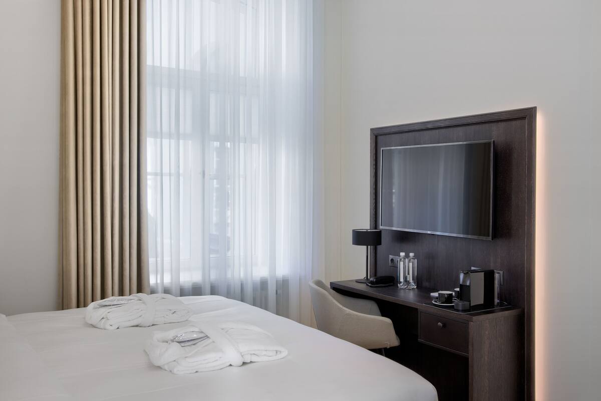 Ein Hotelzimmer mit einem Bett, einem Schreibtisch mit Flachbildfernseher, einem Sessel und einem Fenster mit transparenten Vorhängen.