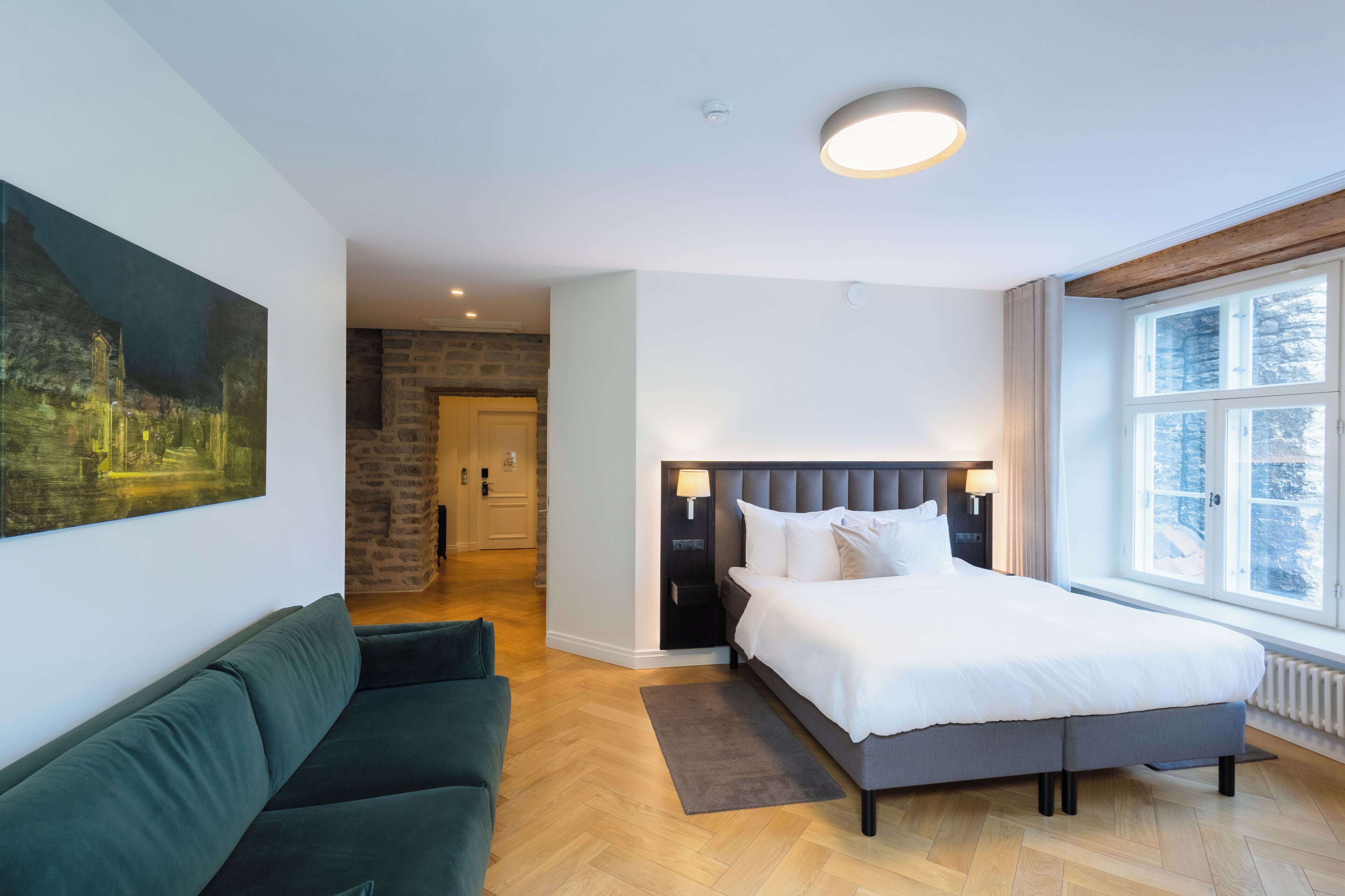 Hotelkamer met een bed, een groene bank, een schilderij aan de muur en uitzicht op bomen door het raam.