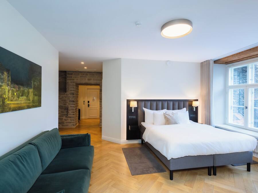 Hotelkamer met een bed, een groene bank, een schilderij aan de muur en uitzicht op bomen door het raam.