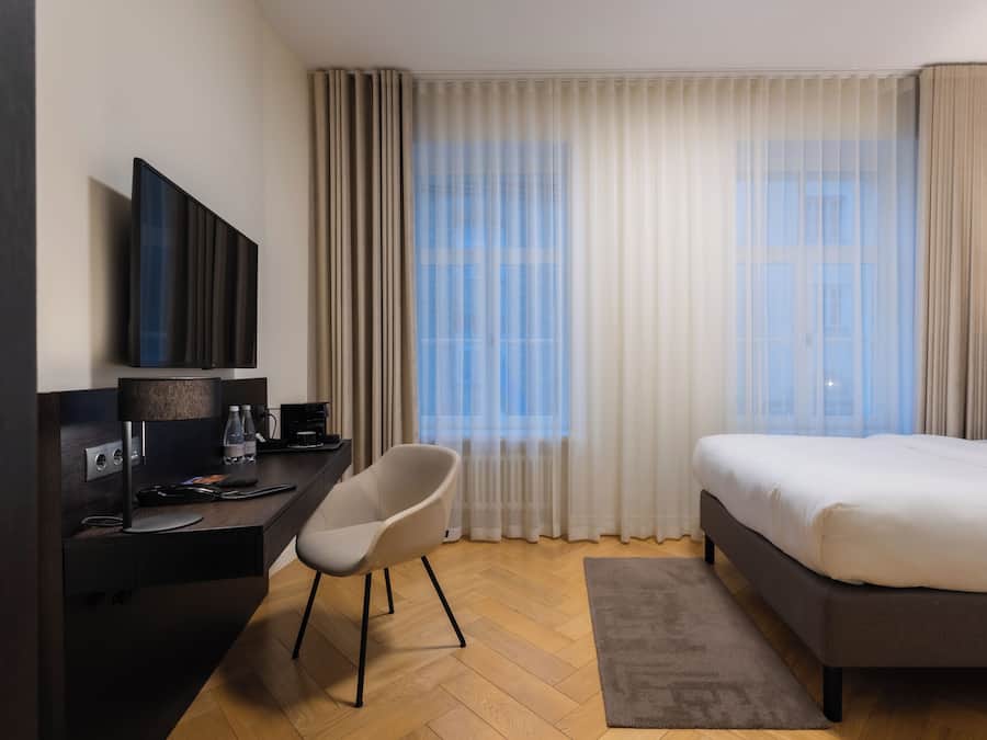 A hotel room with a bed, a desk with a chair, a flat-screen TV, and a large window with sheer curtains.