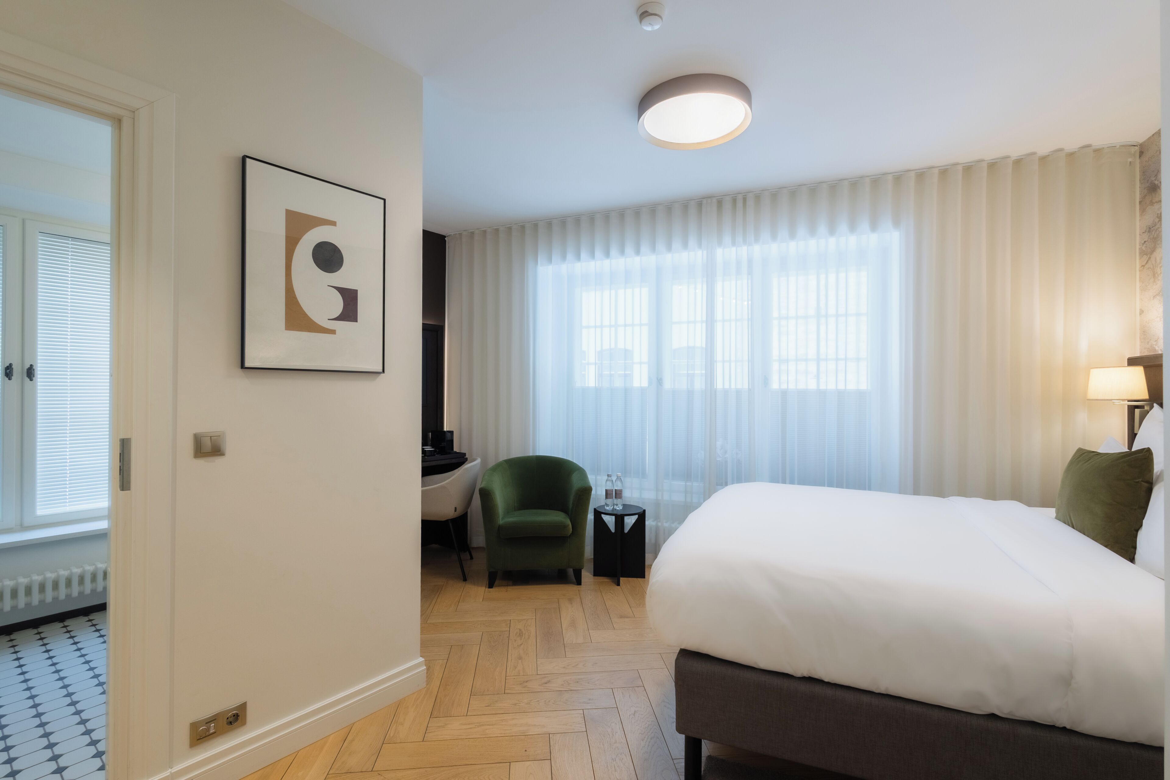 Ein Hotelzimmer mit Bett, Sessel, kleinem Tisch, einem Bild an der Wand und einem Fenster mit Vorhängen.