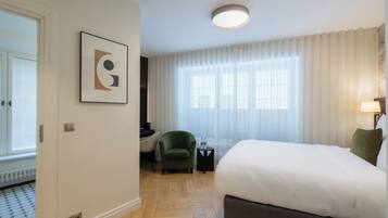 Ein Hotelzimmer mit Bett, Sessel, kleinem Tisch, einem Bild an der Wand und einem Fenster mit Vorhängen.