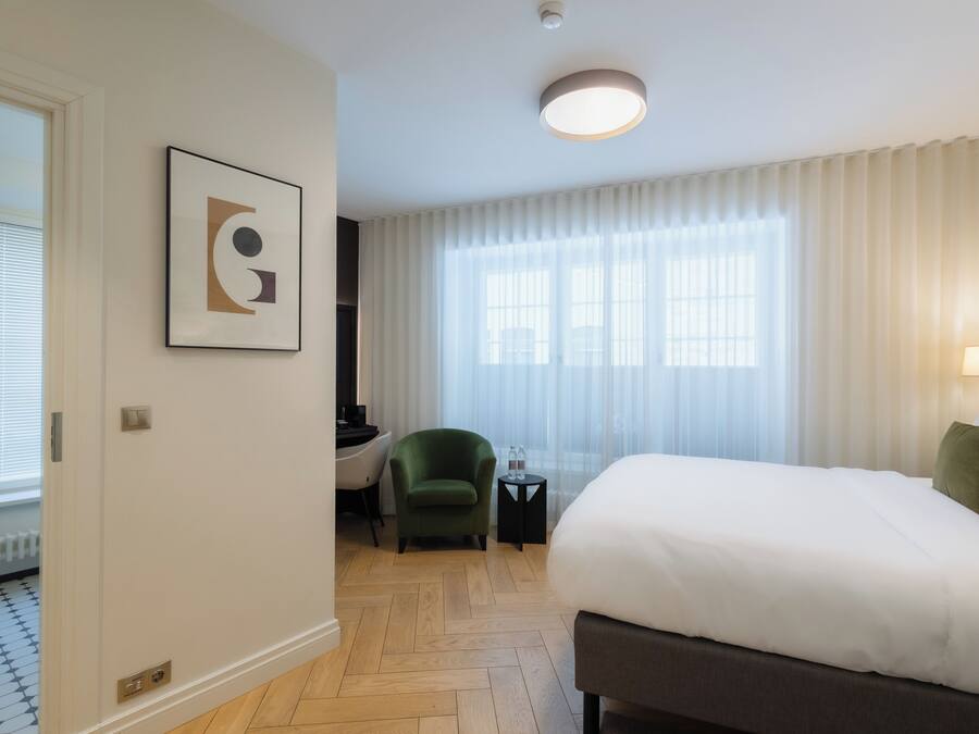 Ein Hotelzimmer mit Bett, Sessel, kleinem Tisch, einem Bild an der Wand und einem Fenster mit Vorhängen.