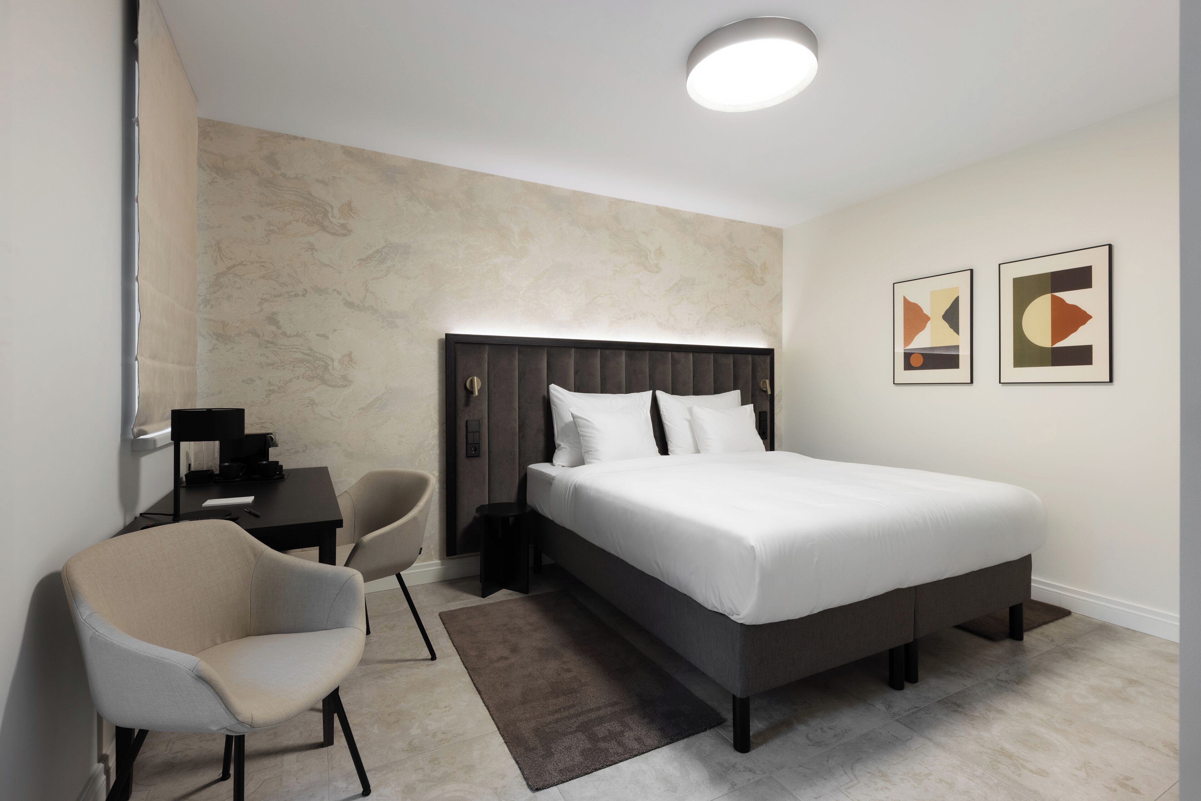 A modern hotel room with a large bed, a desk, a chair, and two abstract paintings on the wall.