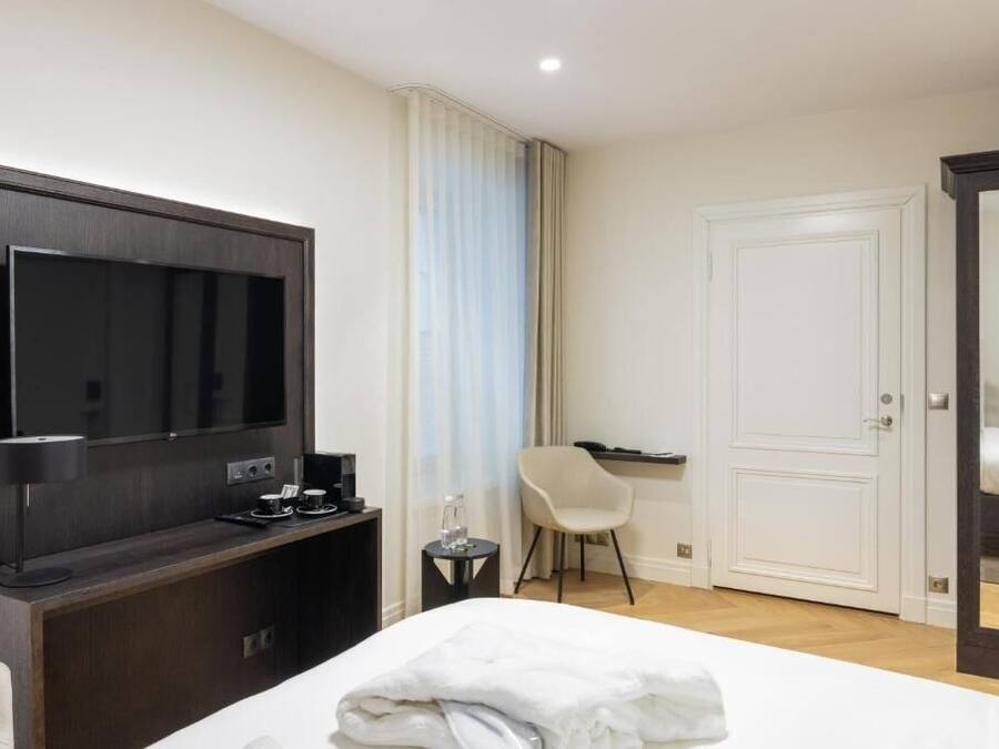 A hotel room with a bed, a TV, a desk with a coffee maker, a chair, and a door.