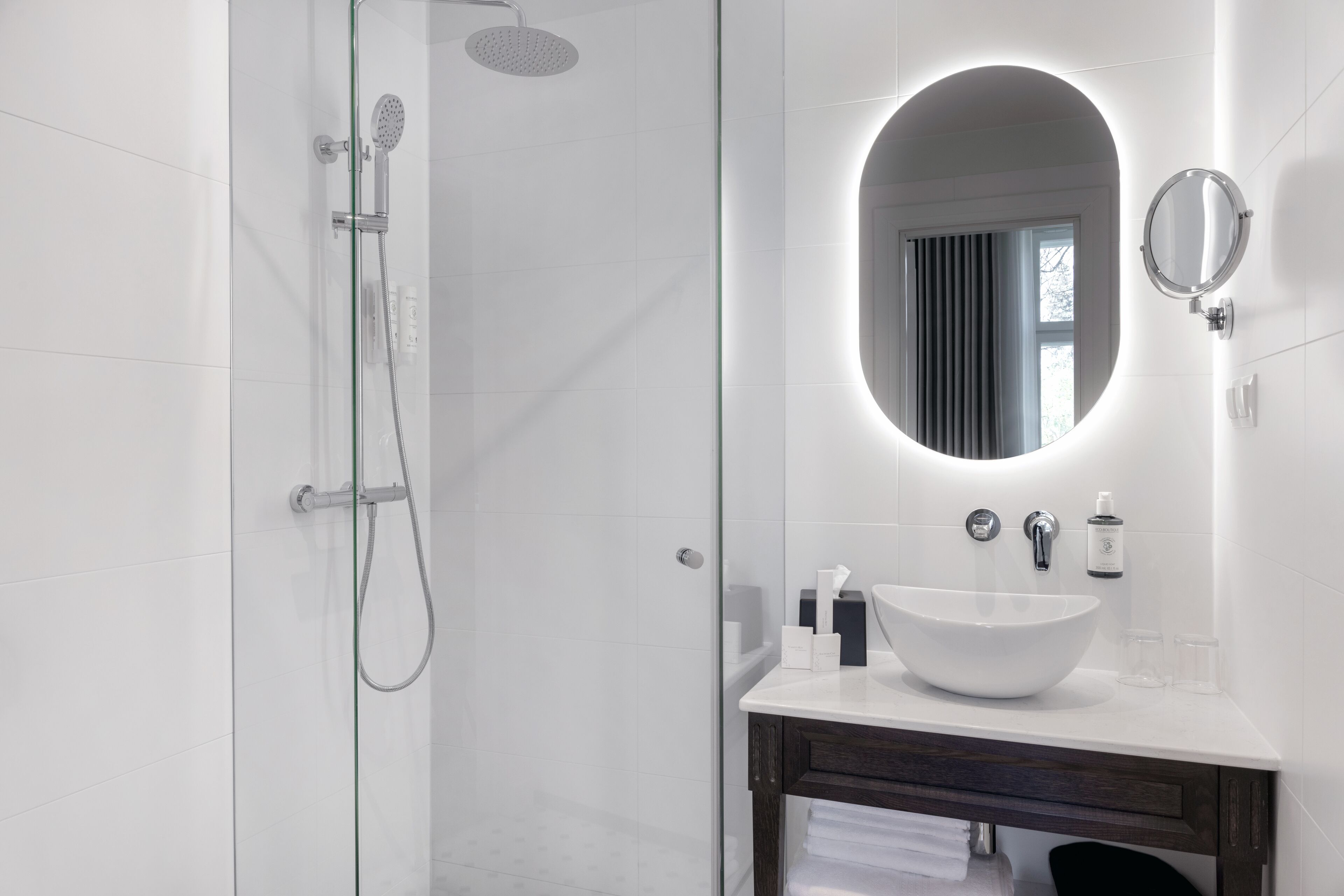 A modern bathroom with a glass shower enclosure, a white sink with a round mirror, and a dark wooden vanity.