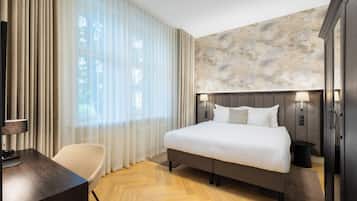 Hotelkamer met een groot bed, een bureau met stoel, een kledingkast en uitzicht op het omliggende landschap door dunne gordijnen.