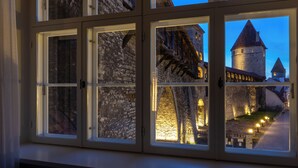 Une vue à travers une fenêtre d’un bâtiment en pierre doté d’une tour, éclairé par une lumière intérieure.