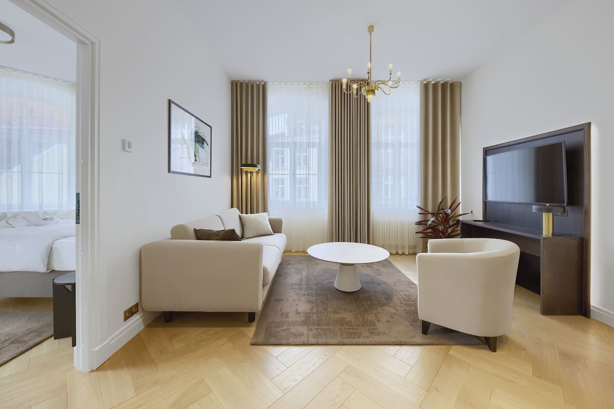 Ein modernes Wohnzimmer mit Sofa, Sessel und Couchtisch, einem Flachbildfernseher und einem großen Fenster mit Vorhängen.