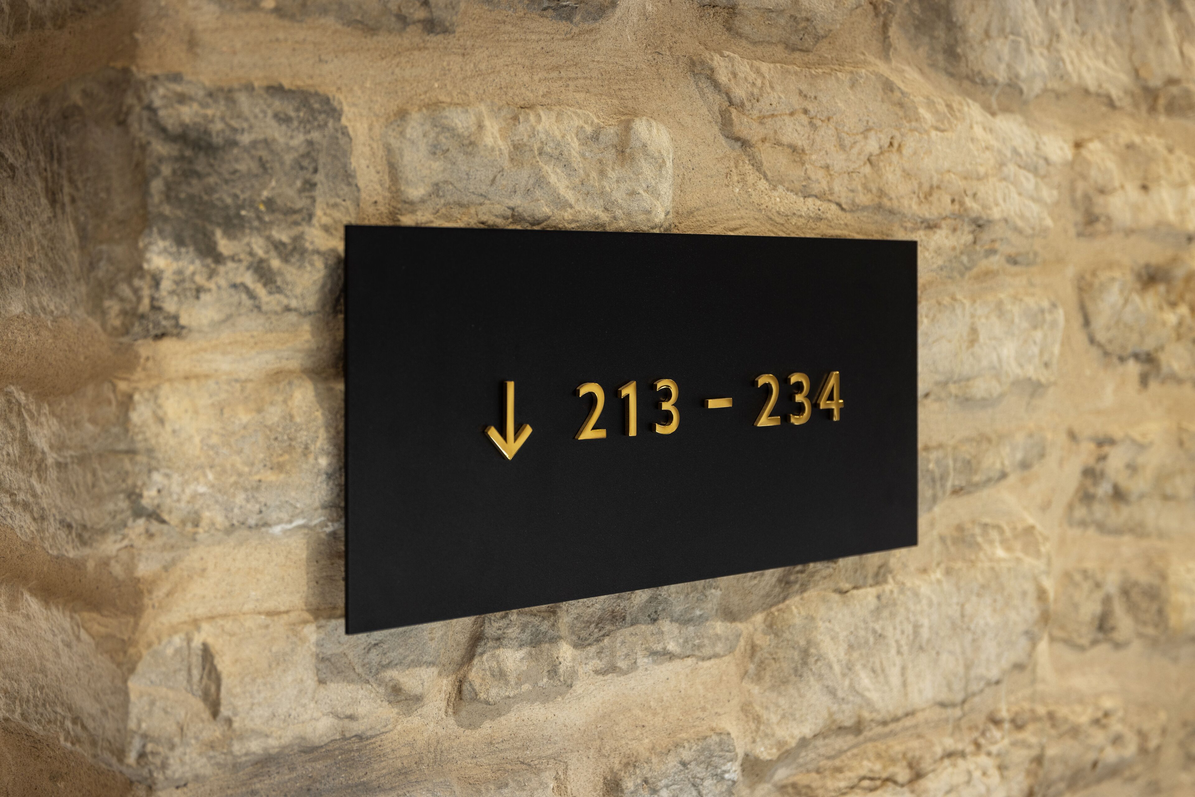 A black sign with gold numbers and an arrow on a stone wall.