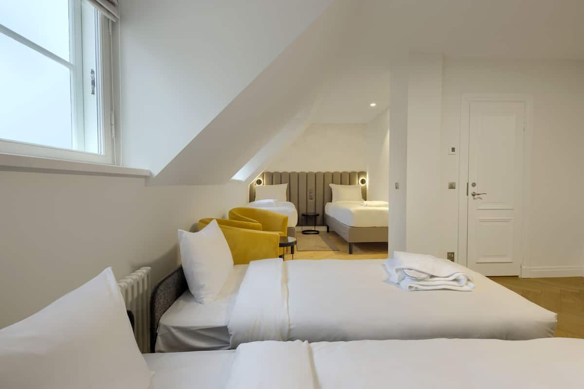 Ein modernes Hotelzimmer mit zwei Betten, einer Sitzecke mit gelben Stühlen und Blick auf die Stadt, sowie einer Tür zum Badezimmer.