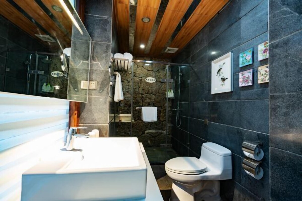 A modern bathroom with a stone wall, a wooden ceiling, a large sink, a toilet, and a glass shower enclosure.