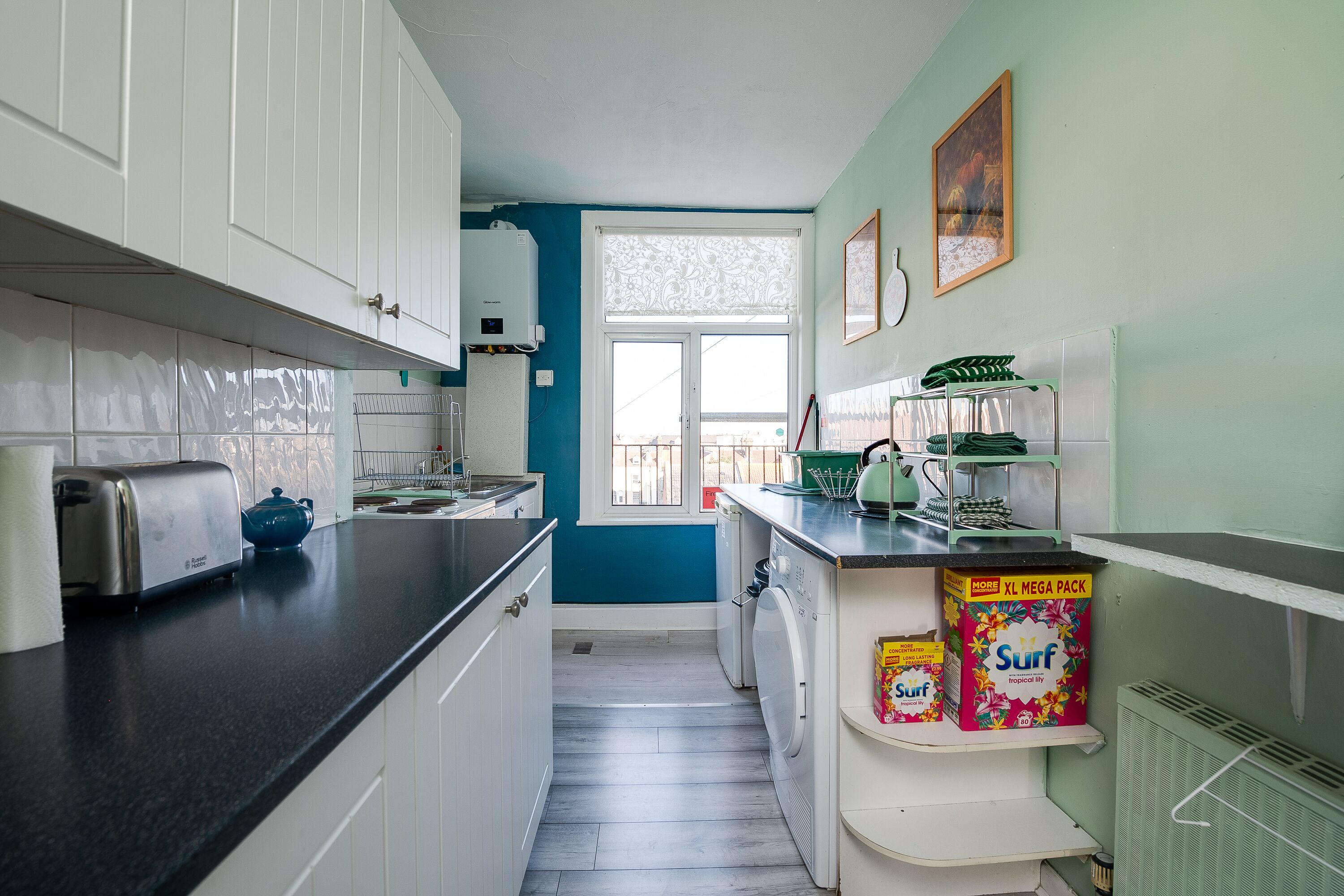 A compact kitchen with white cabinets, a black countertop, a washing machine, and a window with lace curtains.