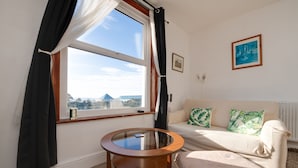 A room with a round glass table, a sofa with patterned cushions, a framed picture on the wall, and a window with curtains.