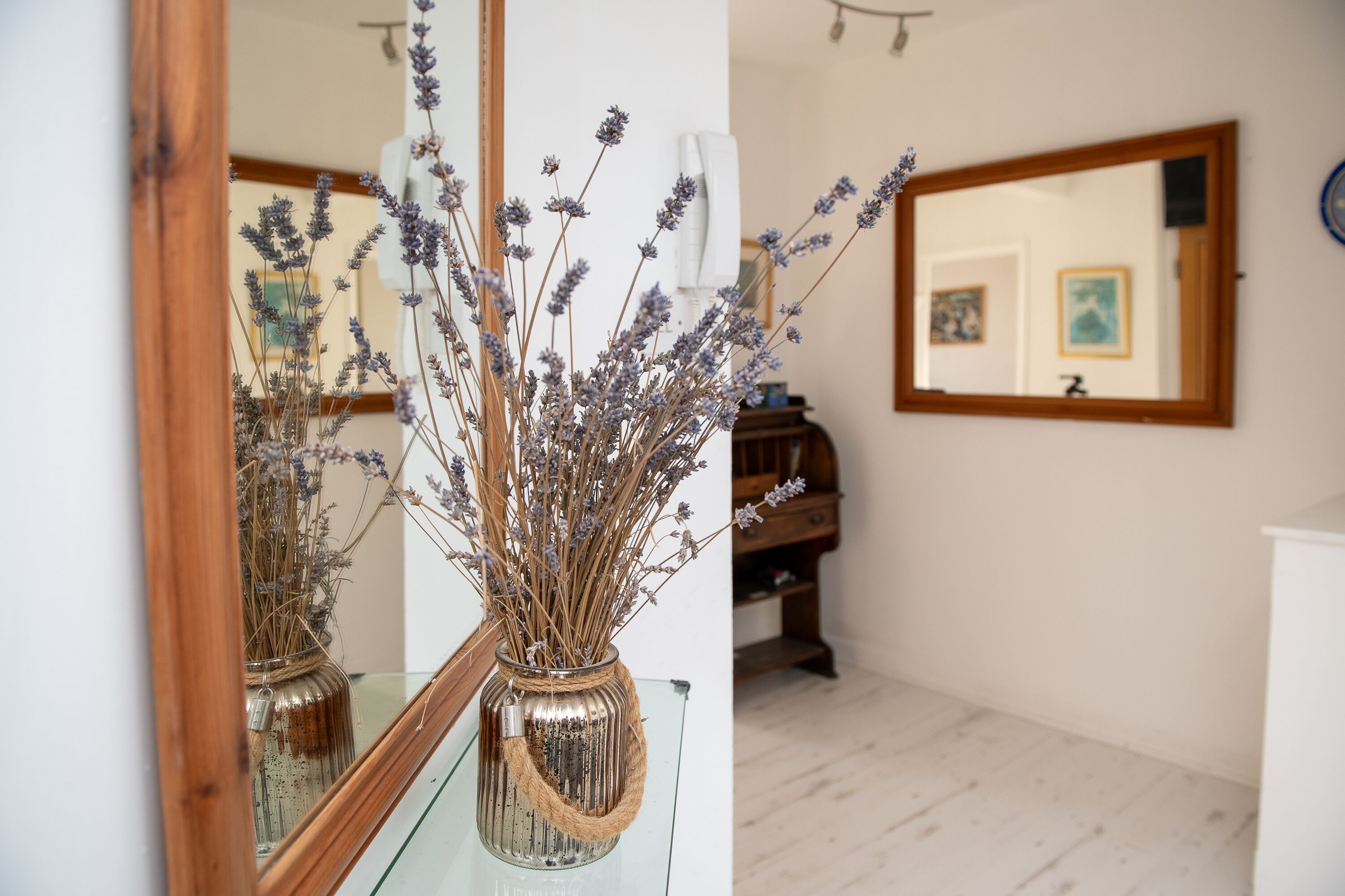 Un miroir reflétant une pièce avec une table en verre, un vase contenant de la lavande séchée et, au fond, un meuble en bois.