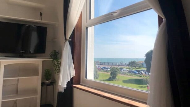A room with a television on a shelf, a window with curtains, and a view of a parking lot and greenery outside.