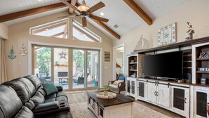A spacious living room with a leather couch, a coffee table, and a TV on a wooden cabinet. There is a large window providing natural light and a view of the outdoors. - Outdoor Sanctuary/Lake View/Hot tub & Game Room! (Canyon Lake)