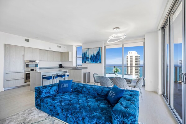 Un apartamento moderno con sala de estar que incluye un sofá azul, una cocina con armarios blancos y un balcón con vista a la ciudad.