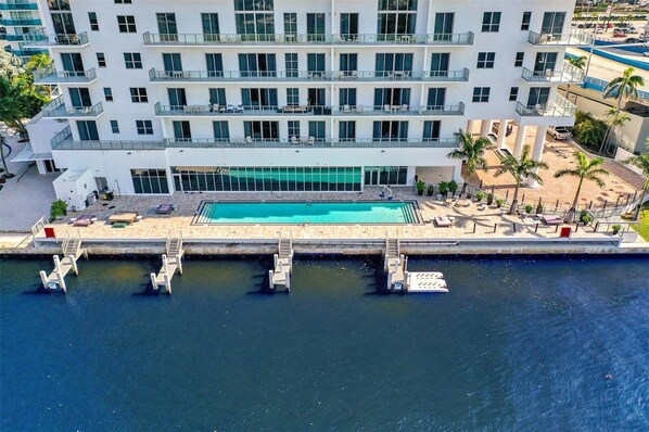 A multi-story hotel with a rooftop pool, a dock with lounge chairs, and a view of the water.