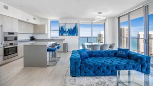 A modern kitchen with stainless steel appliances, a central island, and a dining area with a blue upholstered sofa and a glass coffee table.