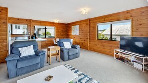 A cozy living room with wooden walls, two blue armchairs, a white coffee table, a TV, and a small kitchen area visible through an open door. - Crystal Clear - Snells Beach Holiday Home (Snells Beach)