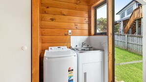 A compact laundry room with a washing machine and a sink, wooden walls, and a window overlooking a backyard. - Crystal Clear - Snells Beach Holiday Home (Snells Beach)