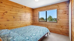 A small, cozy room with wooden walls, a single bed with a patterned bedspread, and a window offering a view of greenery and a clear sky. - Crystal Clear - Snells Beach Holiday Home (Snells Beach)