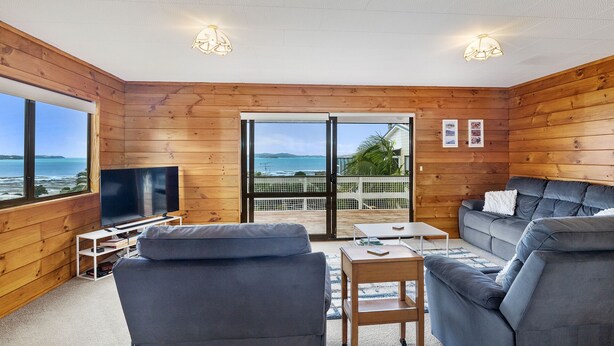 A wooden-paneled room with a TV, grey sofas, and a balcony with a view of the sea. - Crystal Clear - Snells Beach Holiday Home (Snells Beach)