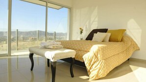 A bedroom with a large window, a bed with a patterned bedspread, a bedside table with towels, and a view of the landscape.